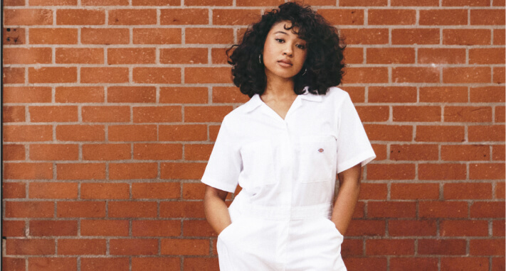 Singer/Dancer Kona standing in front of a brick wall with her hands in her pockets.